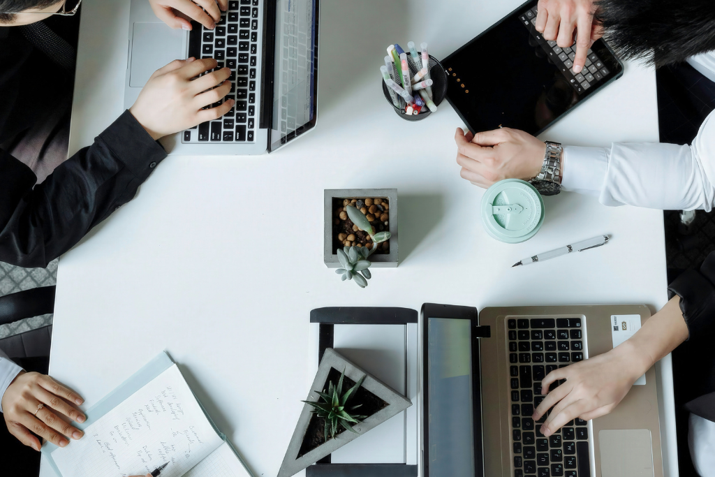 Top view of a collaborative team working on laptops at a modern office desk with notebooks, plants, and work accessories.